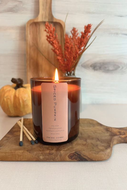 A brown Spiced Pumpkin Candle with a label in front, placed on a wooden tray with autumn-themed decorations in the background.